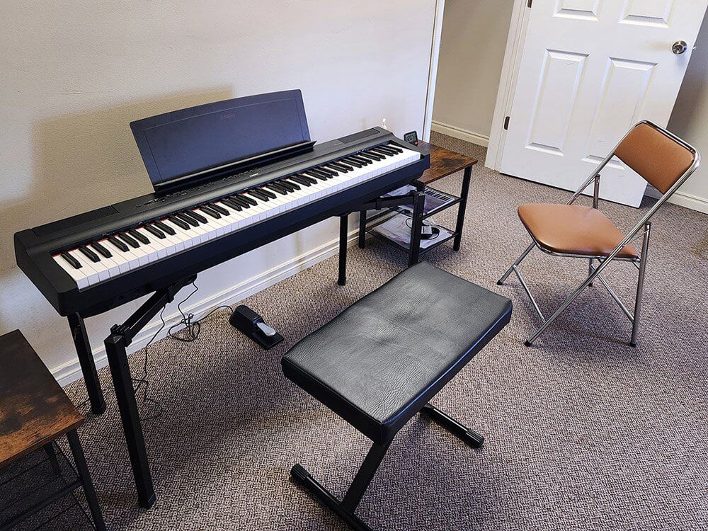 A digital piano with a black stand is placed against a wall in a carpeted room. A black padded bench sits in front of it. A brown folding chair is nearby, and a small side table holds a few items. A white door is in the background.